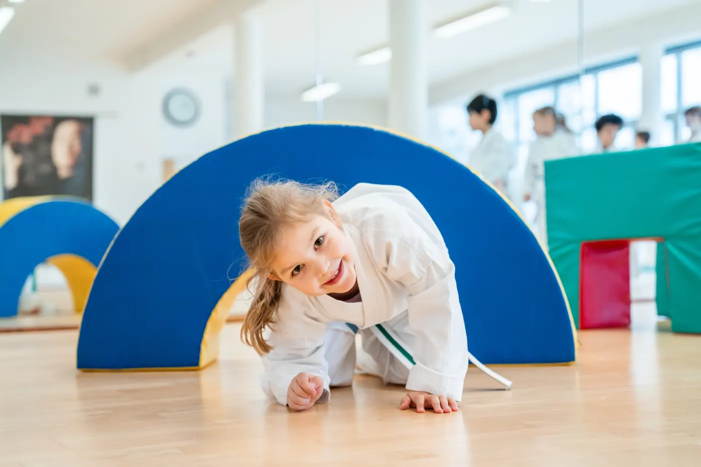 Lächelndes Mädchen im Taekwon-Do-Anzug krabbelt unter einem blauen Bogen hindurch
