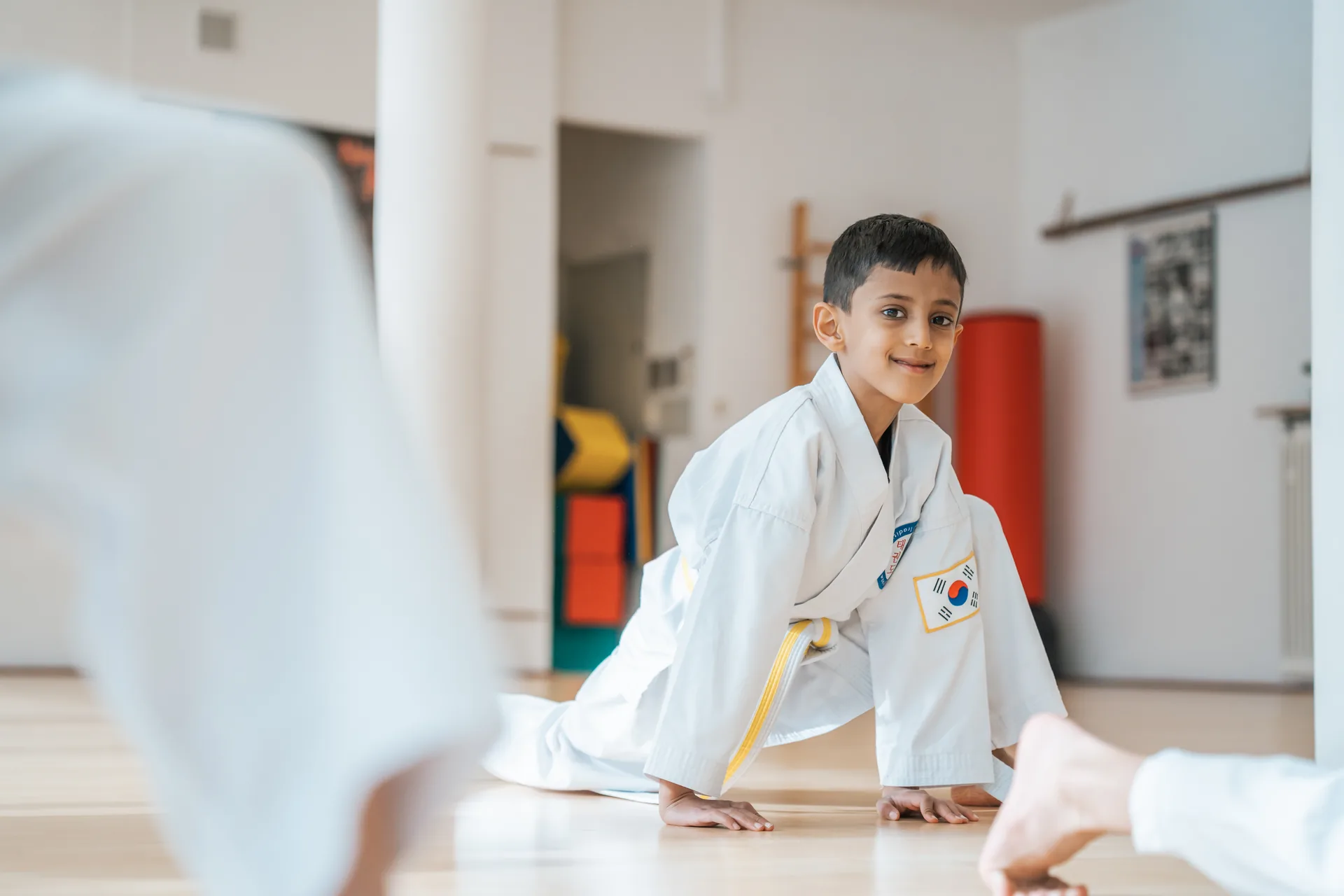 Lächelnder Junge im Taekwon-Do-Anzug mit gelbem Gürtel in tiefer Haltung am Boden, Blick in die Kamera